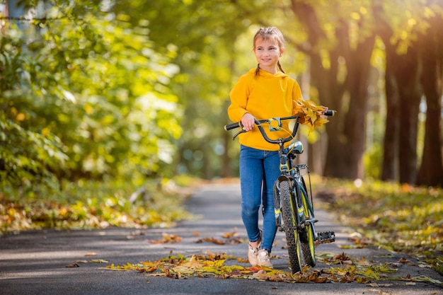 La ragazza sorridente in maglione giallo cammina con la bicicletta in un parco autunnale in una giornata di sole e agita una mano amichevole