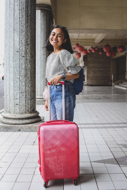 La ragazza sorridente in stile casual porta la valigia passeggiando per la città