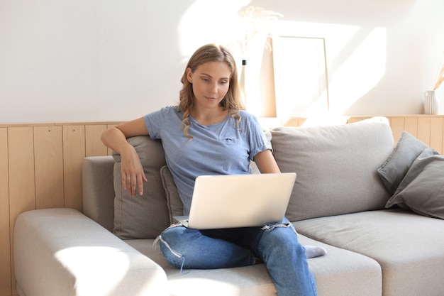 La ragazza sorridente si siede sul divano a guardare il webinar sul computer portatile. Studio felice della giovane donna sul corso a distanza in linea.