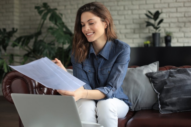 La ragazza sorridente si siede sul divano a guardare il webinar sul computer portatile. Studio felice della giovane donna sul corso a distanza in linea.