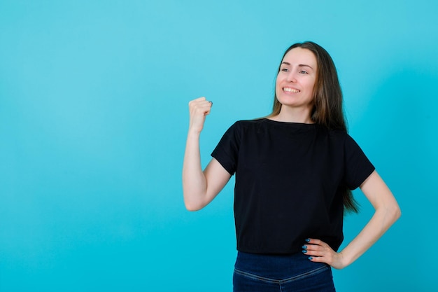 La ragazza sorridente sta alzando il pugno e sta mettendo l'altra mano sulla vita su fondo blu