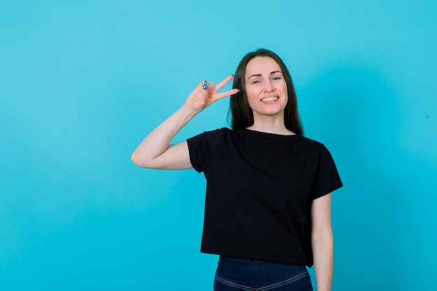 La ragazza sorridente sta mostrando due gesti vicino all'occhio su sfondo blu