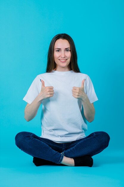 La ragazza sorridente sta mostrando i gesti perfetti sedendosi sul pavimento su fondo blu