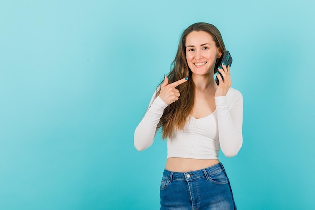 La ragazza sorridente sta mostrando il cellulare in mano con l'indice su sfondo blu