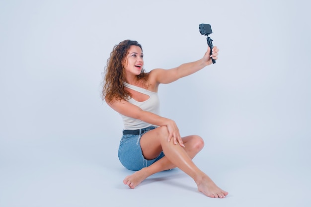 La ragazza sorridente sta prendendo il suo selfie con la sua mini macchina fotografica sedendosi sul pavimento su fondo bianco