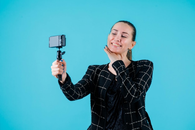 La ragazza sorridente sta prendendo selfie con la sua macchina fotografica tenendo la mano sotto il mento su sfondo blu