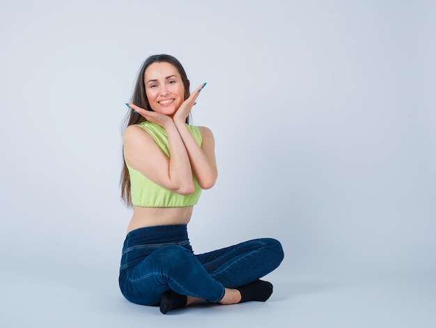 La ragazza sorridente sta tenendo le mani sotto il mento sedendosi sul pavimento su fondo bianco