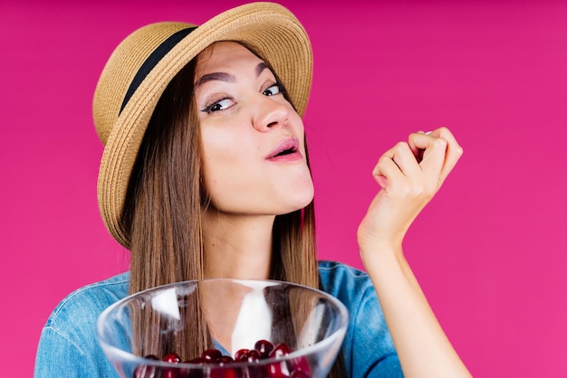 La ragazza sorridente vivace e stupita con un cappello mangia le bacche di ciliegia da un vaso sullo sfondo della sua mano rosa