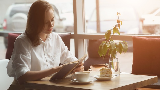 La ragazza sta leggendo un libro seduta in un bar e si gode un caffè e una torta, la luce del sole