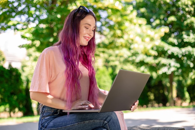 La ragazza sta studiando nel parco primaverile seduta sulla panca di legno e navigando sul suo laptop