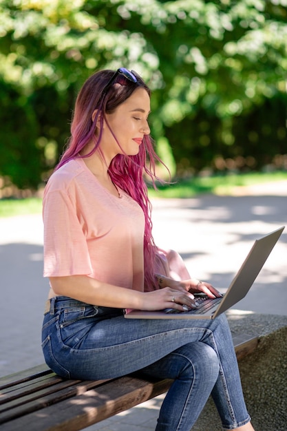 La ragazza sta studiando nel parco primaverile seduta sulla panca di legno e navigando sul suo laptop