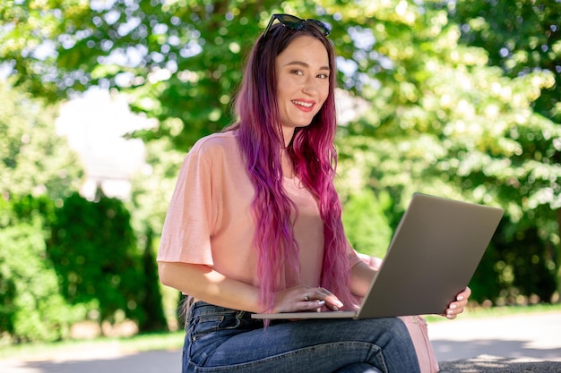 La ragazza sta studiando nel parco primaverile seduta sulla panca di legno e navigando sul suo laptop