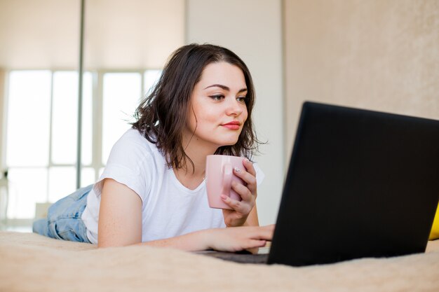 La ragazza sta trovandosi a casa su un letto con caffè e un computer portatile