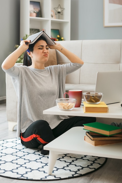 La ragazza stanca ha coperto la testa con il libro che si siede sul pavimento dietro il tavolino da caffè nel soggiorno