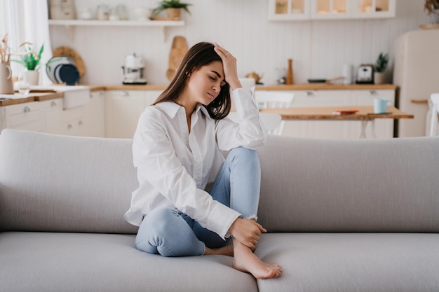 La ragazza stanca seduta sul divano vestita con camicia bianca e jeans blu sente la fatica appoggiarsi sulla mano