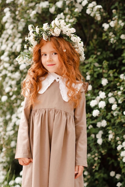 La ragazza sveglia del bambino con i capelli rossi ricci lunghi indossa la corona floreale e il vestito alla moda sopra lo sfondo della natura