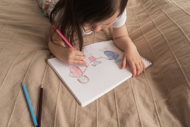 La ragazza sveglia disegna un disegno a matita. Un bambino impara a disegnare le persone. Il bambino si trova sul letto e disegna con una matita