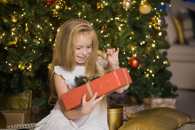 La ragazza sveglia felice si siede vicino all'albero di Natale con il contenitore di regalo rosso