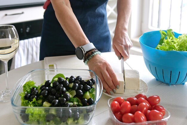 La ragazza taglia il formaggio feta per l'insalata greca preparando il cibo a casa in cucina