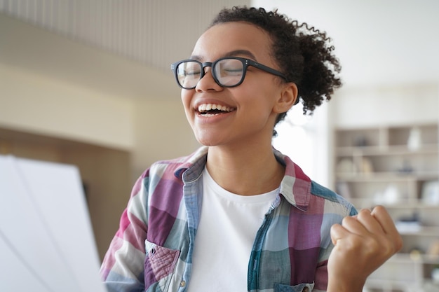 La ragazza teenager afroamericana eccitata con gli occhiali celebra la vittoria e gode del successo personale