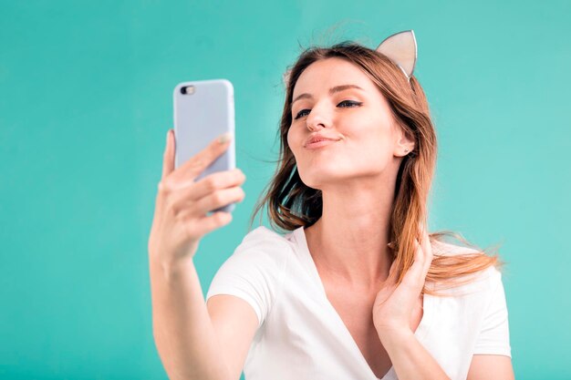 La ragazza teenager bionda sorridente felice fa il selfie del bacio tramite smartphone su fondo blu