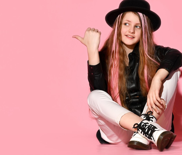La ragazza teenager mostra il dito nella direzione dello spazio libero seduto sul pavimento con una giacca di pelle e un cappello con un bordo