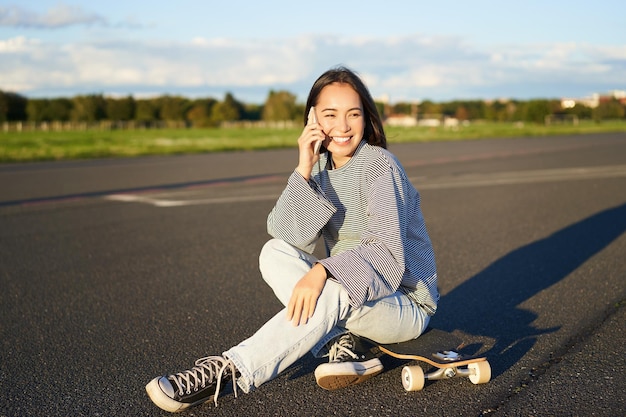 La ragazza teenager sveglia si siede sullo skateboard e parla sulla donna felice del pattinatore del telefono cellulare che ha conversazione o