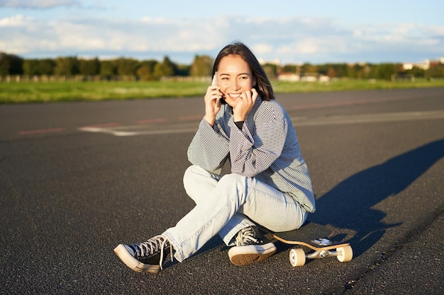 La ragazza teenager sveglia si siede sullo skateboard e parla sulla donna felice del pattinatore del telefono cellulare che ha conversazione o