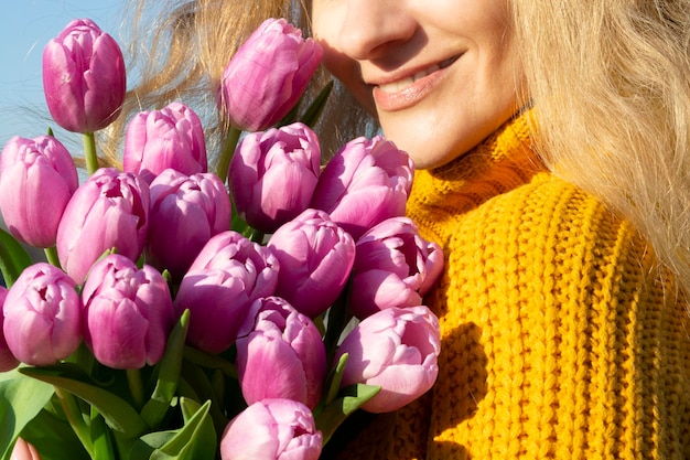 La ragazza tiene i tulipani accanto al viso fiori viola una ragazza con un maglione giallo in maglia appoggiò la guancia contro i fiori sullo sfondo di un cielo blu