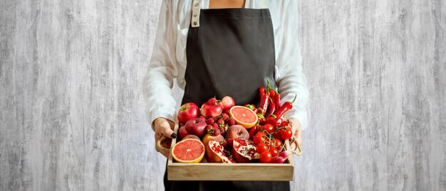 La ragazza tiene il vassoio di legno con le verdure e la frutta rosse fresche su fondo grigio. Concetto vegetariano di cibo sano.