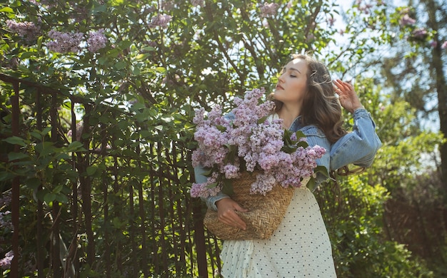 La ragazza tiene tra le mani un cesto di vimini con fiori. Cesto con lillà. Ragazza e fiori. Cammina con un cesto di lillà in mano. Floristica. Fiore di lillà primaverile