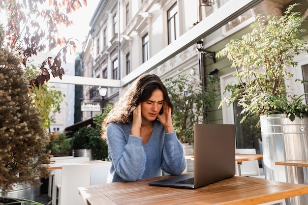 La ragazza triste con il laptop ha avuto l'emicrania le tocca la testa a causa del dolore Donna manager con forte mal di testa che lavora sul laptop online da remoto all'aperto nel caffè