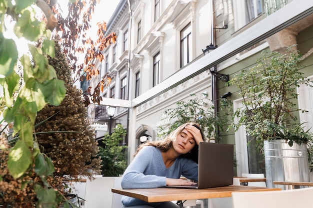 La ragazza triste con il laptop ha avuto l'emicrania le tocca la testa a causa del dolore Donna manager con forte mal di testa che lavora sul laptop online da remoto all'aperto nel caffè