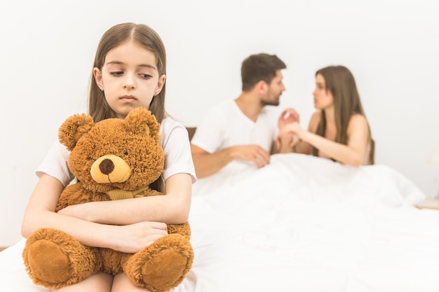 La ragazza triste con un giocattolo si siede vicino ai genitori sul letto