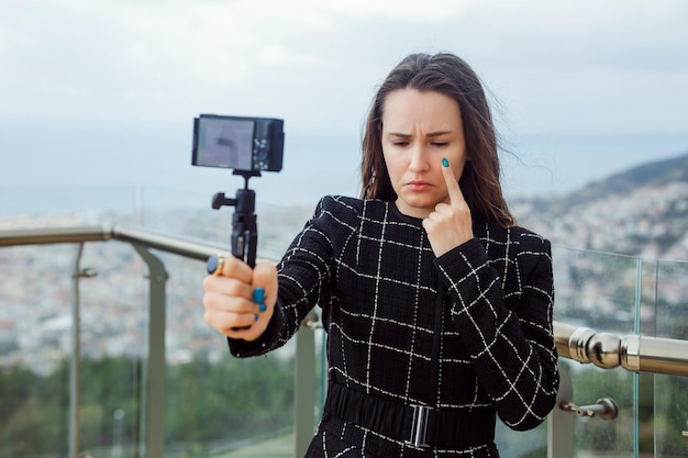 La ragazza triste del blogger si sta facendo un selfie con la sua mini fotocamera tenendo l'indice sulla guancia sullo sfondo della vista della città