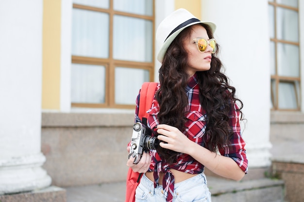 La ragazza turista con cappello e occhiali da sole che tiene la telecamera contro l'edificio con colonne