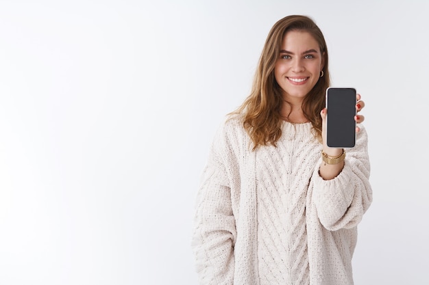 La ragazza vuole sentire la tua opinione mostrandoti il display dello smartphone. Attraente e affascinante donna dall'aspetto amichevole che estende la mano che tiene il telefono cellulare condividendo un'immagine divertente che sorride ampiamente sfondo bianco