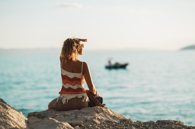 La retrovisione di una giovane donna attraente sta godendo di una vacanza estiva mentre si siede e guarda sul mare.