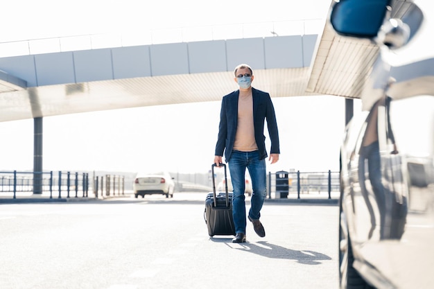 La ripresa all'aperto di un uomo adulto torna dal viaggio di lavoro va in auto con la valigia indossa una maschera medica durante l'epidemia di coronavirus La malattia pandemica Il passeggero arriva dopo un viaggio internazionale