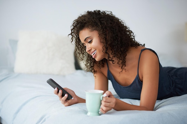 La routine mattutina per controllare i social media Inquadratura di una giovane donna attraente sdraiata sul letto e che usa il cellulare mentre tiene una tazza di caffè