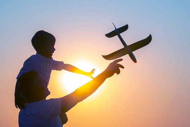 La sagoma di un ragazzo, seduto sulla spalla di suo padre, lancia un aeroplano modello nel cielo della sera sullo sfondo del sole al tramonto. Il sogno dei bambini di un futuro pilota