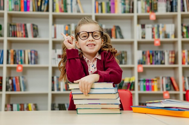 La scolara con gli occhiali è seduta a una scrivania con un'enorme pila di libri di testo in biblioteca