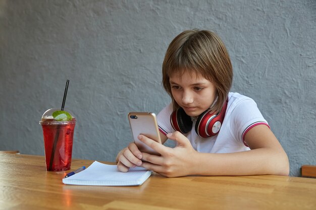 La scolara con le cuffie e la limonata ghiacciata rossa utilizza lo smartphone naviga in internet