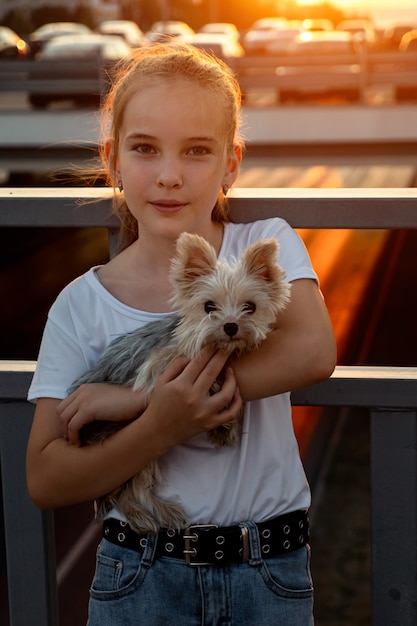 La scolara posa con il cane al tramonto posteriore. L'adolescente in maglietta bianca tiene in braccio un piccolo cucciolo in piedi sul ponte di strada contro la luce del sole al tramonto