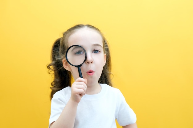 La scolaretta curiosa positiva in abiti casual guarda la telecamera attraverso una lente d'ingrandimento su uno sfondo giallo sorpresa