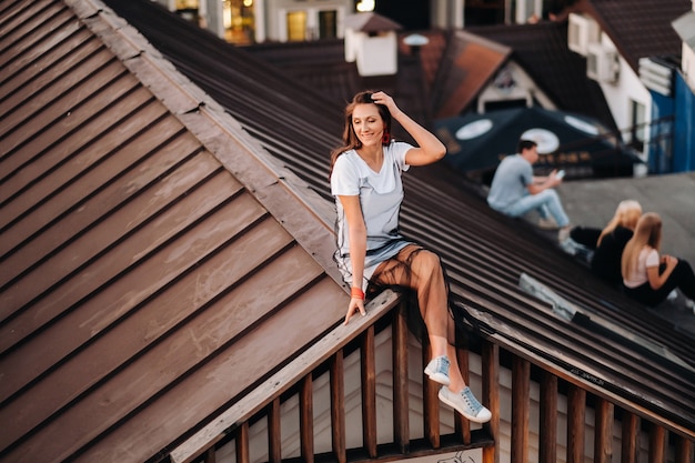 La sera una ragazza si siede sul tetto di una casa in città. Ritratto di un modello in un vestito e scarpe da ginnastica.