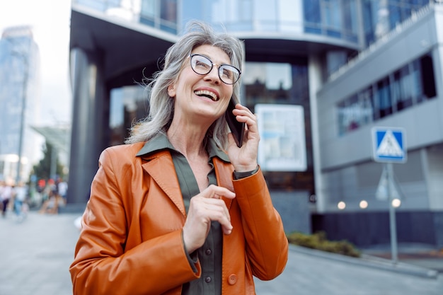 La signora anziana dai capelli canuto allegra parla al cellulare sulla strada moderna della città