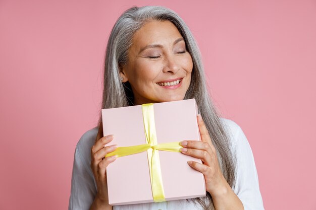 La signora asiatica matura allegra tiene una scatola regalo con un nastro in posa su sfondo rosa