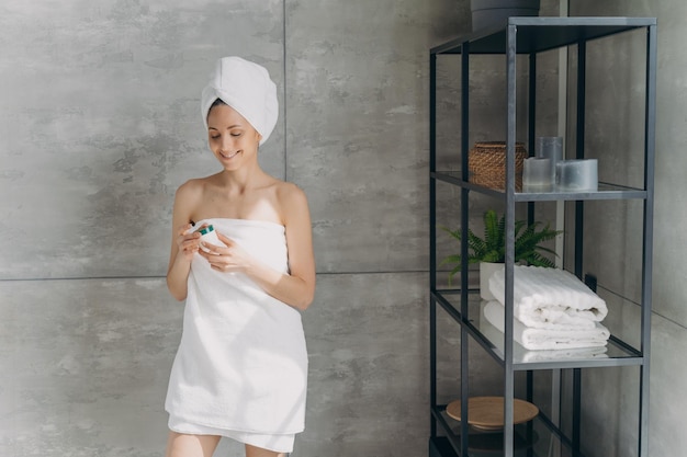La signora europea applica la crema Giovane donna felice avvolta in un asciugamano dopo il bagno e il lavaggio dei capelli