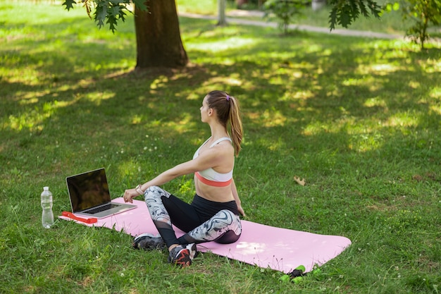 La signora flessibile fa una torsione del corpo seduta vicino a un laptop aperto su un tappetino rosa nel parco estivo